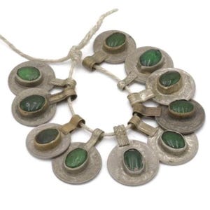 May include: A necklace featuring multiple silver-toned, circular pendants with a central, oval-shaped, green stone. The pendants are connected by a white cord. The design has a vintage or antique aesthetic.