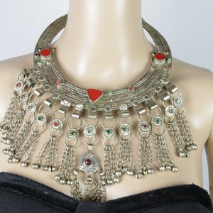 Può includere: Una collana d'argento con un grande ciondolo a forma di mezzaluna ornato. Il ciondolo è decorato con pietre rosse e verdi e ha una frangia di catene d'argento con piccoli campanellini.