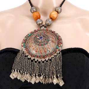 May include: An elaborate silver necklace with a large, decorative pendant. The pendant is adorned with colourful gemstones, detailed metalwork, and chain tassels. The necklace includes amber beads and a black cord.