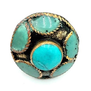 May include: A round, ornate ring featuring a central turquoise stone surrounded by smaller turquoise accents. The ring has a dark base with gold-colored metal accents and a textured design, creating a vintage or bohemian aesthetic.