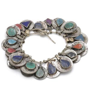 May include: A silver-toned necklace featuring coin-shaped pendants with colorful gemstone accents. The gemstones are various shapes and colors, including turquoise, red, blue, and purple. The necklace is strung on a natural-colored cord.