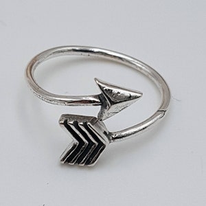 May include: A silver-colored ring with an arrow design. The arrow features a triangular head and a series of chevron-shaped lines. The ring is adjustable and sits on a white surface.
