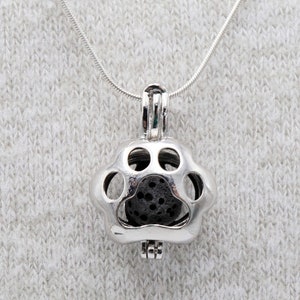 May include: A silver-colored paw print pendant necklace. The pendant is shaped like a paw with open circles and contains a dark, porous stone. The necklace chain is thin and silver, set against a light gray background.