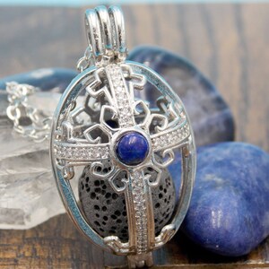 May include: Silver oval pendant with a cross design and a blue gemstone in the centre. The pendant is attached to a silver chain and is surrounded by smaller stones.