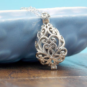 May include: A silver-toned metal pendant necklace featuring an intricate, openwork filigree design shaped like a teardrop. The pendant is suspended from a delicate silver-colored chain, showcasing detailed swirling patterns and a central floral motif.