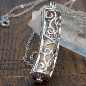 Könnte beinhalten: Eine silberne Halskette mit einem zarten, verzierten Anhänger. Der Anhänger ist ein hohler Zylinder mit einem filigranen Design und einem weißen, strukturierten Material im Inneren. Die Halskette liegt auf einer Holzoberfläche.