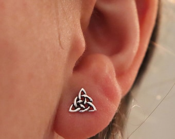 May include: Silver Celtic knot earrings in the shape of a triangle. The earrings are small and delicate, and they are shown in a person's ear.
