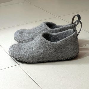 Puede incluir: Un par de zapatillas de lana afieltrada gris con punta redondeada, abertura baja y una lengüeta de cuero gris oscuro en el talón. Las zapatillas tienen un patrón de punto de cruz visible a lo largo de la base donde la parte superior se une a la suela.