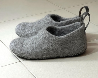 May include: A pair of gray felted wool slippers featuring a rounded toe, a low-cut opening, and a dark gray leather pull tab at the heel. The slippers have a visible stitched seam along the base where the upper meets the sole.