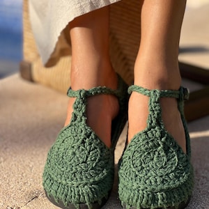 May include: A pair of dark green crochet sandals featuring a circular pattern on the toe, a T-strap design, and a buckled ankle strap. The footwear has a solid black sole and is constructed with thick, textured yarn in a deep forest green shade.