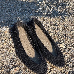 May include: A pair of black espadrille flats with a woven upper and a natural-colored jute sole. The shoes are displayed on a bed of small pebbles, showcasing their casual, summery style. The flats are designed for comfort and breathability.