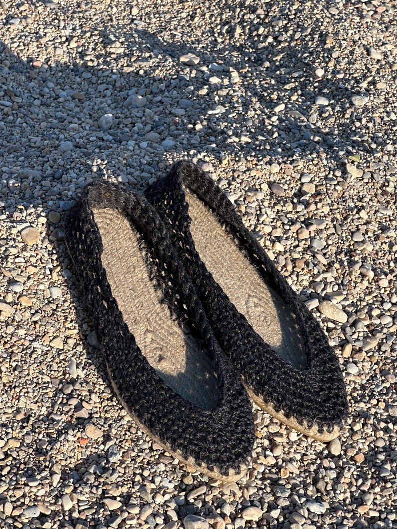 May include: A pair of black espadrille flats with a woven upper and a natural-colored jute sole. The shoes are displayed on a bed of small pebbles, showcasing their casual, summery style. The flats are designed for comfort and breathability.