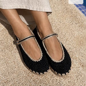 May include: Black crochet Mary Jane flats featuring a textured knit upper, a beige braided trim along the collar, and a matching beige braided strap with a small button closure. The shoes have a thick, light-colored jute-style rope sole.
