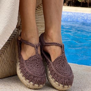 May include: A pair of brown crochet shoes featuring a T-strap design with a small wooden button closure. The upper part is crafted from dark brown yarn in a circular pattern, while the base consists of a thick, braided jute rope sole.