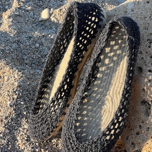 May include: A pair of black espadrille flats with a woven, open-weave design. The shoes have a natural-colored jute sole and are resting on a sandy surface with small pebbles and a rock.