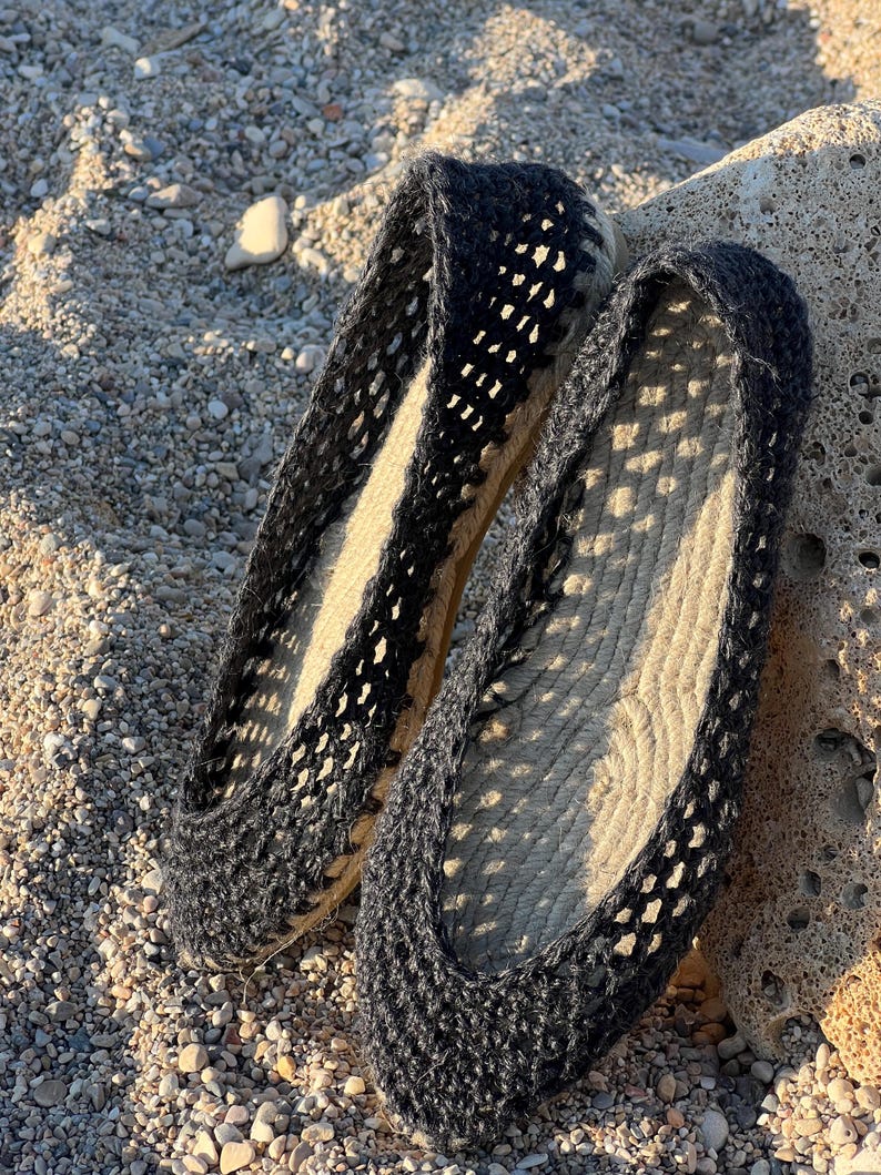 May include: A pair of black espadrille flats with a woven, open-weave design. The shoes have a natural-colored jute sole and are resting on a sandy surface with small pebbles and a rock.