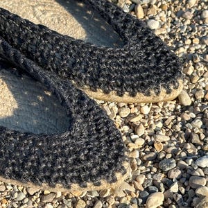 May include: Close-up of black espadrille shoes. The shoes have a woven jute sole and a black knitted upper. The shoes are resting on a bed of small pebbles, suggesting an outdoor setting. The design is simple and casual.