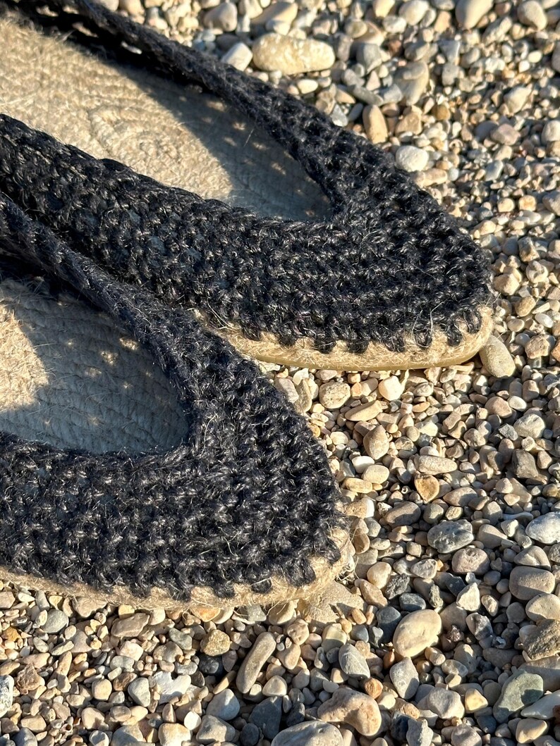 May include: Close-up of black espadrille shoes. The shoes have a woven jute sole and a black knitted upper. The shoes are resting on a bed of small pebbles, suggesting an outdoor setting. The design is simple and casual.