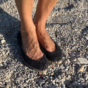 May include: Black crochet flats with a rounded toe. The shoes are made of a dark, textured material and have a woven sole. The flats are worn on a pebbled surface.