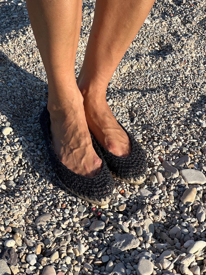 May include: Black crochet flats with a rounded toe. The shoes are made of a dark, textured material and have a woven sole. The flats are worn on a pebbled surface.