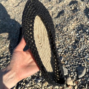 May include: A black crochet espadrille shoe with a natural fiber sole. The shoe is held up, showcasing the open weave of the crochet and the texture of the sole. The shoe is designed for warm weather.