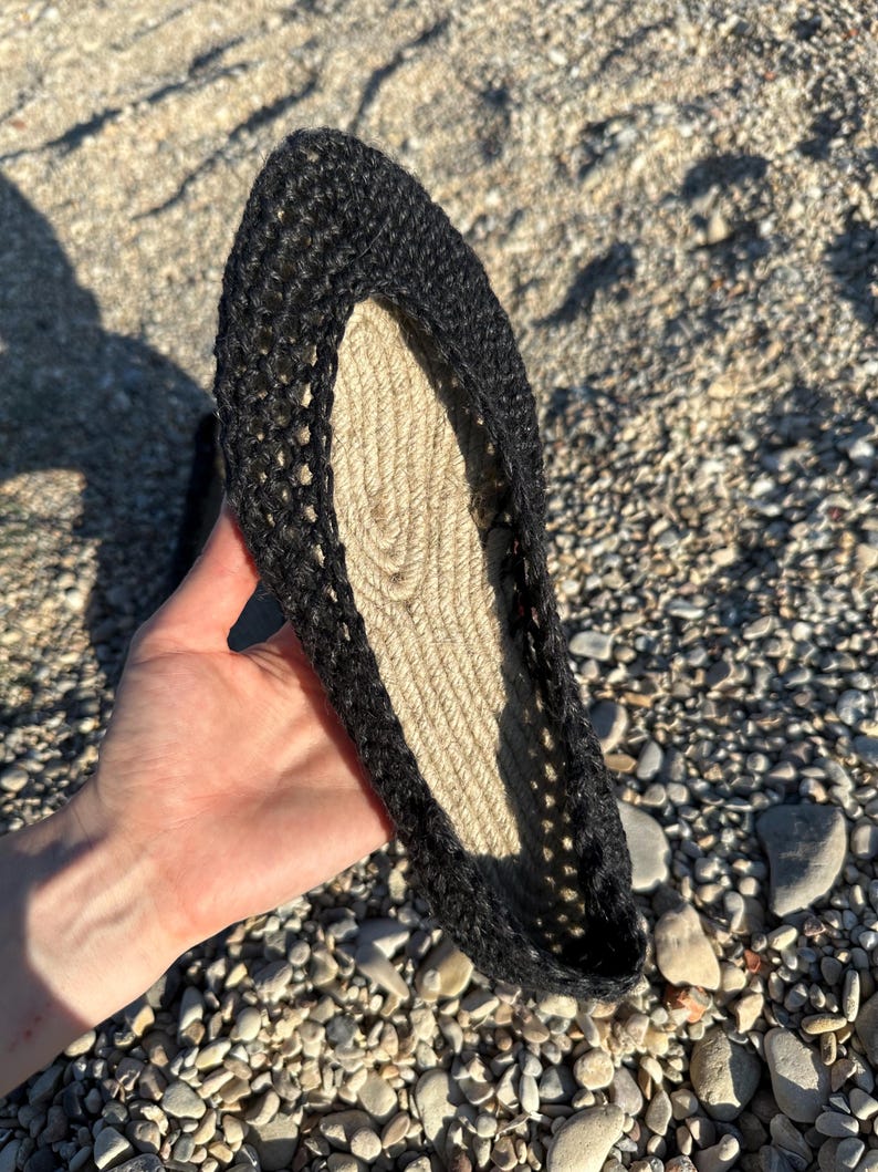 May include: A black crochet espadrille shoe with a natural fiber sole. The shoe is held up, showcasing the open weave of the crochet and the texture of the sole. The shoe is designed for warm weather.