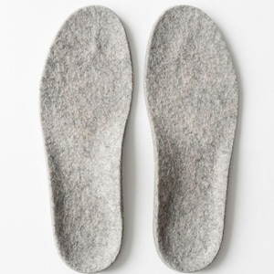May include: A pair of light gray felt insoles with a textured, fibrous surface. The insoles are shaped to fit the contours of a shoe, featuring a rounded heel and a tapered toe area, displaying a consistent mottled gray color throughout the material.