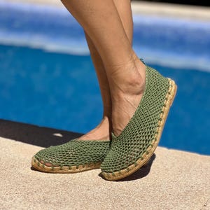May include: A pair of olive green crochet slip-on shoes featuring an open-knit mesh upper design and a natural tan jute rope sole. The footwear has a rounded toe and a low-cut profile, showcasing a textured, woven construction throughout the body.