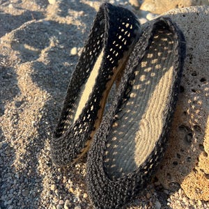 May include: A pair of black espadrille flats with a woven design. The shoes have a rope sole and a dark upper with a cut-out pattern. The flats are resting on a sandy surface with small pebbles and a rock.