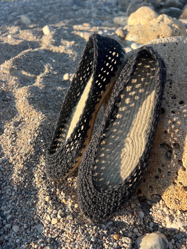 May include: A pair of black espadrille flats with a woven design. The shoes have a rope sole and a dark upper with a cut-out pattern. The flats are resting on a sandy surface with small pebbles and a rock.
