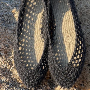 May include: A pair of navy blue espadrille flats. The shoes have a woven jute sole and a dark blue crocheted upper with a perforated design. The shoes are displayed on a pebbled surface.