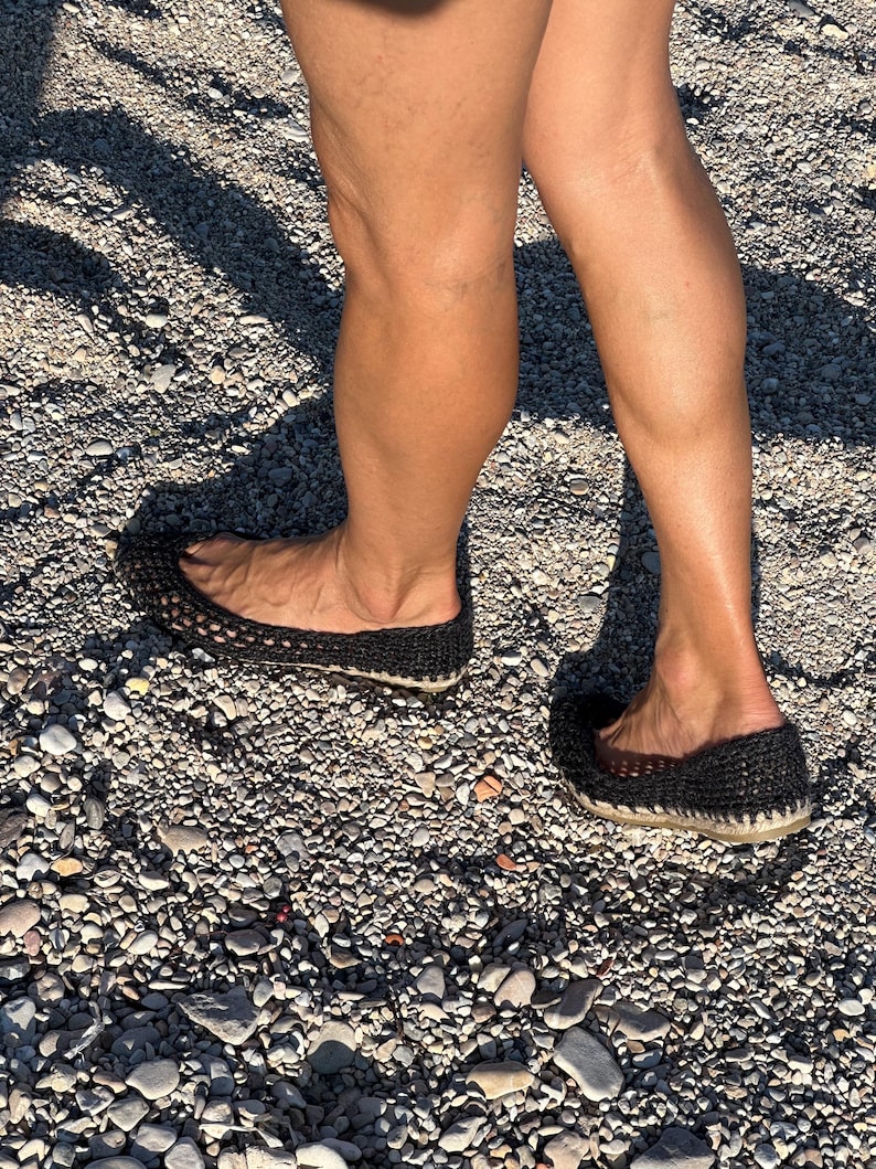 May include: Black crochet flats with a woven design and a light-colored sole. The shoes are worn on a pebbled surface, suggesting outdoor use. The flats appear comfortable and casual, suitable for everyday wear.