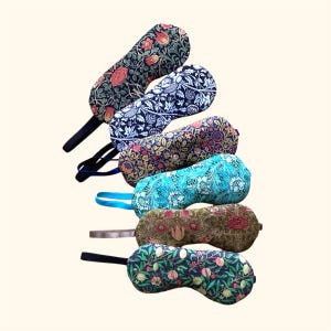 May include: Six sleep masks with floral patterns in a variety of colours. The masks are curved and made of fabric. Each mask features a different design, including red, blue, and green floral prints. The masks have black or coloured elastic straps.