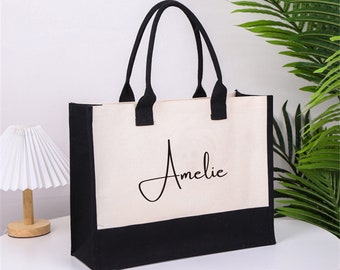Könnte beinhalten: Eine weiße Canvas-Tragetasche mit schwarzem Besatz und Griffen. Die Tasche trägt den Namen "Amelie" in schwarzer Kursivschrift auf der Vorderseite.