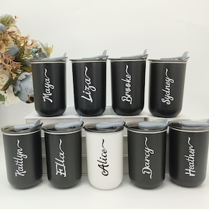Puede incluir: Ocho tazas de viaje personalizadas. Siete son negras y una blanca. Cada taza tiene una tapa gris con cierre deslizante. Los nombres en escritura blanca son: Maya, Liza, Brooke, Sydney, Kaitlyn, Ella, Alice, Darcy y Heather.