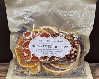 May include: A clear plastic bag filled with dried citrus fruit slices, rose buds, chamomile flowers, lemongrass, and lavender buds. The bag has a label that reads "Bright, Floral, Citrus Kissed SUN-KISSED HOLLOW orange slices, grapefruit slices, rose buds, chamomile flowers, lemongrass, lavender buds, salt".