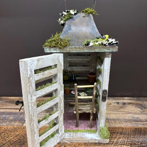 Może przedstawiać: Miniaturowy drewniany szopa z dachem pokrytym mchem i małymi drzwiami, które są otwarte. Wewnątrz szopy znajduje się mały stolik i krzesło, a także kilka miniaturowych roślin i butelek.