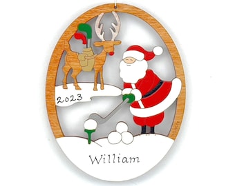 Può includere: Ornamento natalizio in legno raffigurante Babbo Natale che gioca a golf con una renna. L'ornamento ha una cornice in legno marrone chiaro e un interno bianco. L'anno "2023" è visibile, insieme al nome "William".