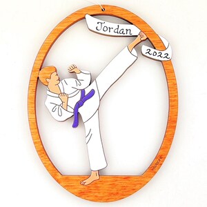 May include: A wooden oval ornament featuring a cartoon illustration of a person in a white karate uniform performing a kick. The ornament has a purple belt and a banner with the name "Jordan" and the year "2022".