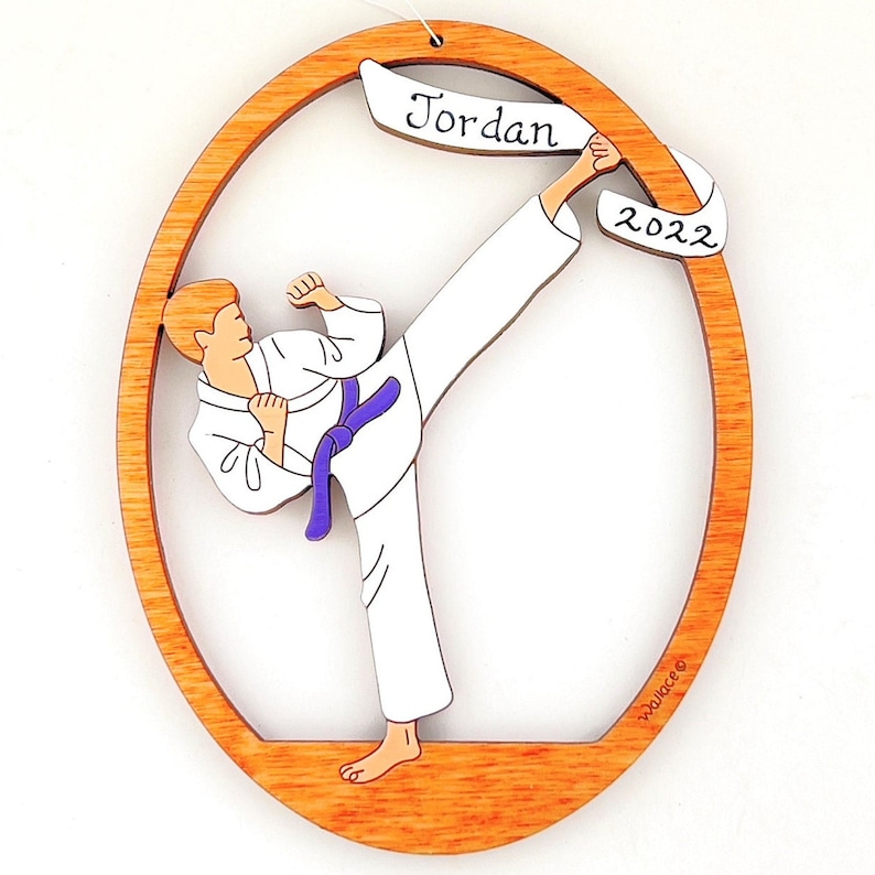 May include: A wooden oval ornament featuring a cartoon illustration of a person in a white karate uniform performing a kick. The ornament has a purple belt and a banner with the name "Jordan" and the year "2022".