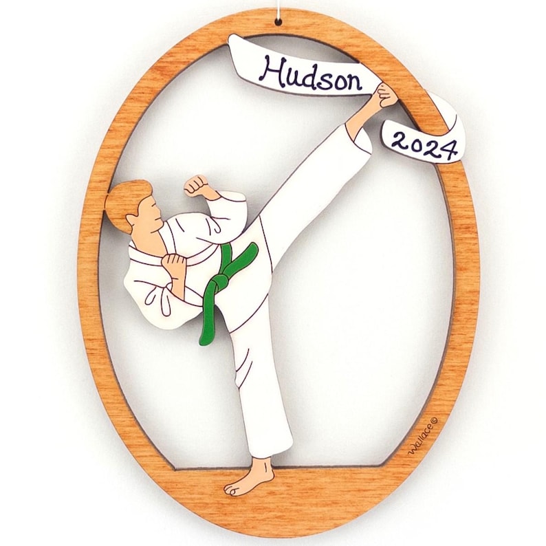 May include: A wooden Christmas ornament featuring a cartoon of a person in a white karate uniform with a green belt, kicking in the air. The ornament is oval shaped with a banner that reads "Hudson 2024".