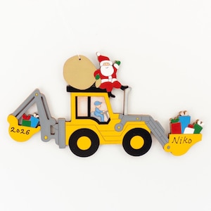 May include: A yellow and gray wooden ornament shaped like a construction vehicle. Santa Claus sits on top, and a driver is inside. The ornament has the year "2026" and the name "Niko" on it, with gifts in the front and back.