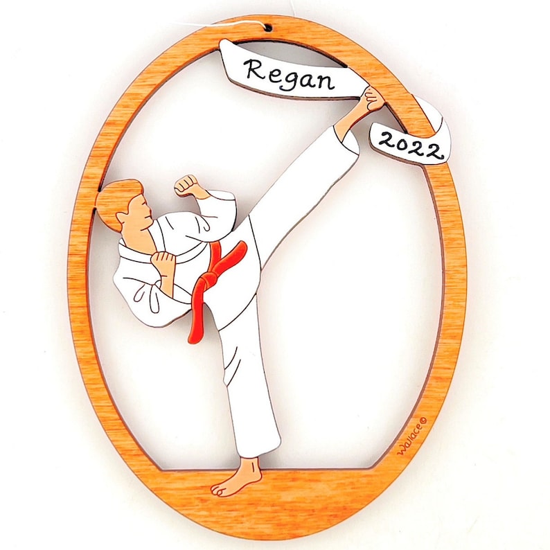 May include: A wooden oval ornament featuring a cartoon illustration of a person in a white karate uniform performing a kick. The ornament has a banner with the name "Regan" and the year "2022" written on it.