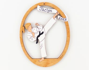 May include: An oval wooden ornament featuring a cutout of a person in a white karate uniform with a black belt performing a high kick. A white banner across the top displays the name Jaclyn and the year 2025 in black script lettering.
