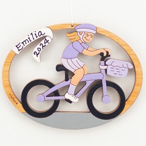 Könnte beinhalten: Ein ovaler Holzanhänger mit einem Mädchen, das ein lila Fahrrad fährt. Das Mädchen trägt einen lila Helm und ein lila Shirt. Der Anhänger hat ein weißes Banner mit dem Text "Emilia 2024".