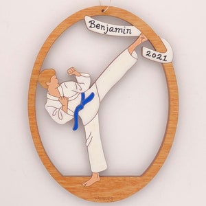 May include: A wooden ornament featuring a cartoon of a person in a white karate uniform with a blue belt, kicking with their right leg. The ornament is oval shaped with a brown frame and a white banner at the top that says "Benjamin 2021".