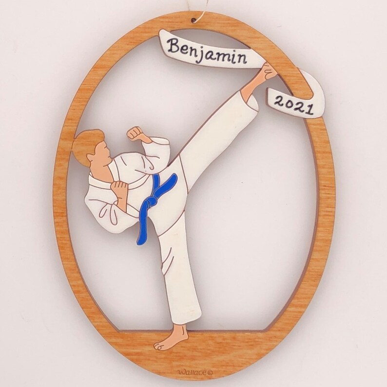 May include: A wooden ornament featuring a cartoon of a person in a white karate uniform with a blue belt, kicking with their right leg. The ornament is oval shaped with a brown frame and a white banner at the top that says "Benjamin 2021".
