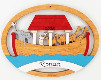 May include: An oval wooden ornament featuring a layered Noah's Ark design with a red roof marked 2026, a figure holding a bird, two elephants, two camels, and two giraffes. A white banner at the bottom displays the name Ronan in purple script.
