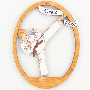 May include: A wooden oval ornament featuring a cartoon illustration of a person in a white karate uniform performing a high kick. The ornament has a white banner with the name "Drew" and the year "2024" written on it.