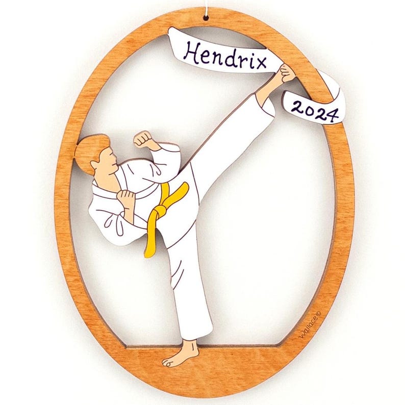 May include: A wooden Christmas ornament featuring a cartoon illustration of a person in a white karate uniform performing a high kick. The ornament is oval shaped with a banner at the top that reads "Hendrix 2024".